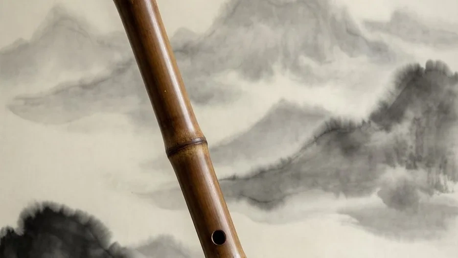 A long vertical xiao end-blown bamboo flute leaning against a stone beside a window, soft evening light falling across its finger holes